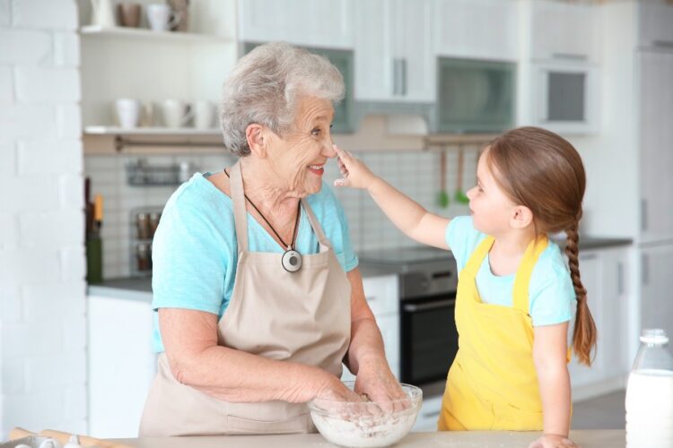 An elderly woman wearing an apron and a medical alert device smiles and interact with a young girl