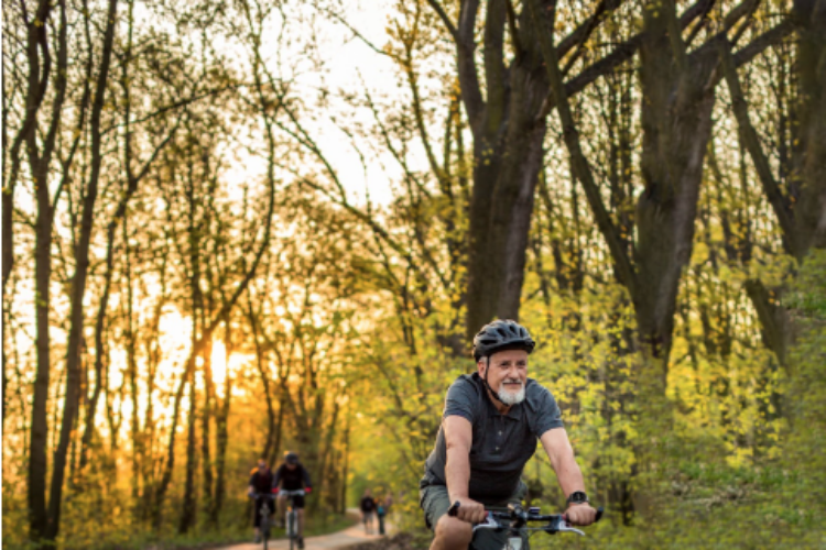 An older man riding a bike on a forest trail during sunset wearing a sidekick mobile medical alert system