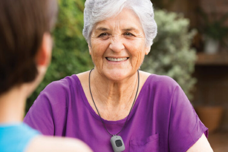 Smiling senior woman wearing LifeStation medical alert pendant, sitting outdoors with companion.